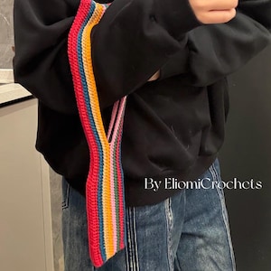 Könnte beinhalten: Ein handgefertigter Häkchen-Taschenriemen mit einem lebendigen Regenbogenmuster in Rot, Blau, Gelb und Grün. Der Riemen wird vor einem schwarzen Sweatshirt und einer blauen Jeans gehalten. Der Text auf dem Bild lautet "By EliomiCrochets".