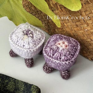 May include: Two handmade crochet turtle-shaped items in shades of purple and lavender. Each turtle has a square body, a white border, and small, dark brown legs. The text "By EliomiCrochets" is visible in the background.