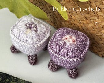 May include: Two handmade crochet turtle-shaped items in shades of purple and lavender. Each turtle has a square body, a white border, and small, dark brown legs. The text "By EliomiCrochets" is visible in the background.