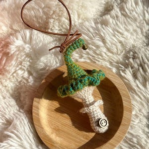 May include: A handmade crochet mushroom ornament with a green and teal cap, a white stem, and a wooden button detail. It has a brown leather loop for hanging and sits on a wooden plate.