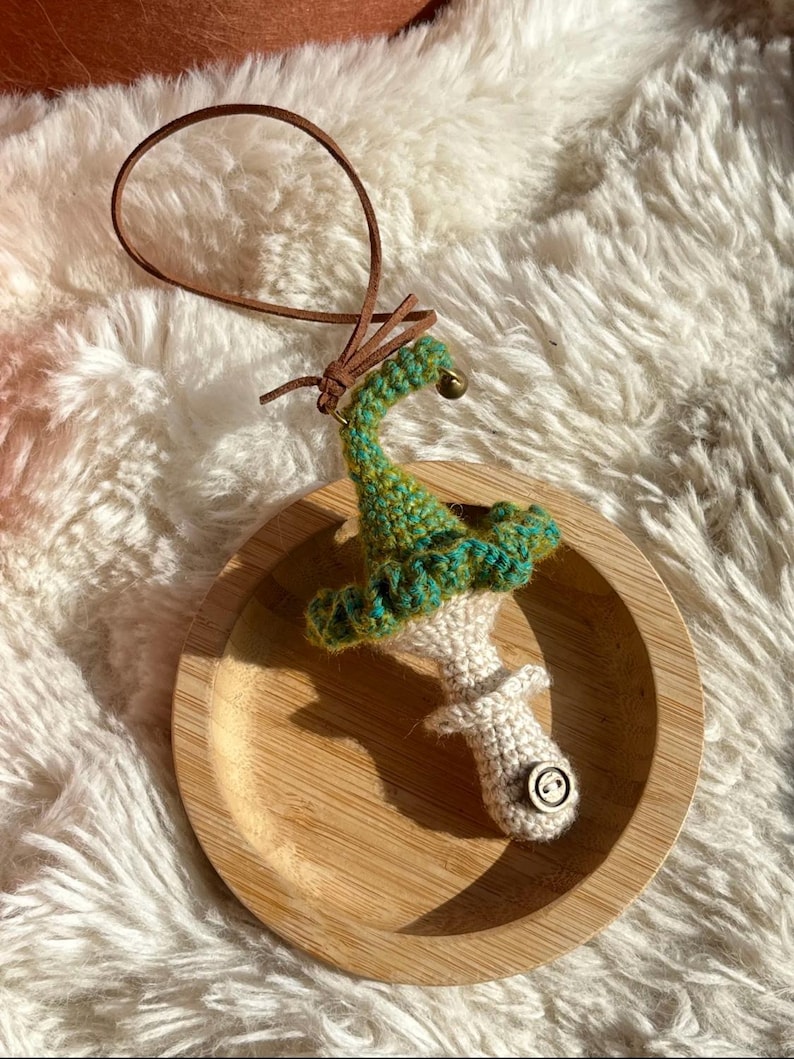 May include: A handmade crochet mushroom ornament with a green and teal cap, a white stem, and a wooden button detail. It has a brown leather loop for hanging and sits on a wooden plate.