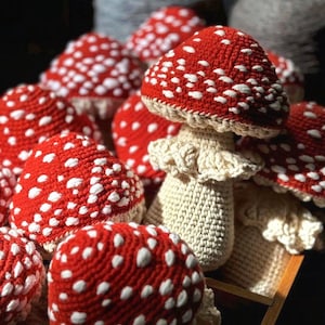 May include: A collection of handmade crochet mushrooms. The mushroom caps are red with white spots, and the stems are off-white. The mushrooms are arranged in a wooden tray, with some in the background. The yarn used appears to be a soft, textured material.