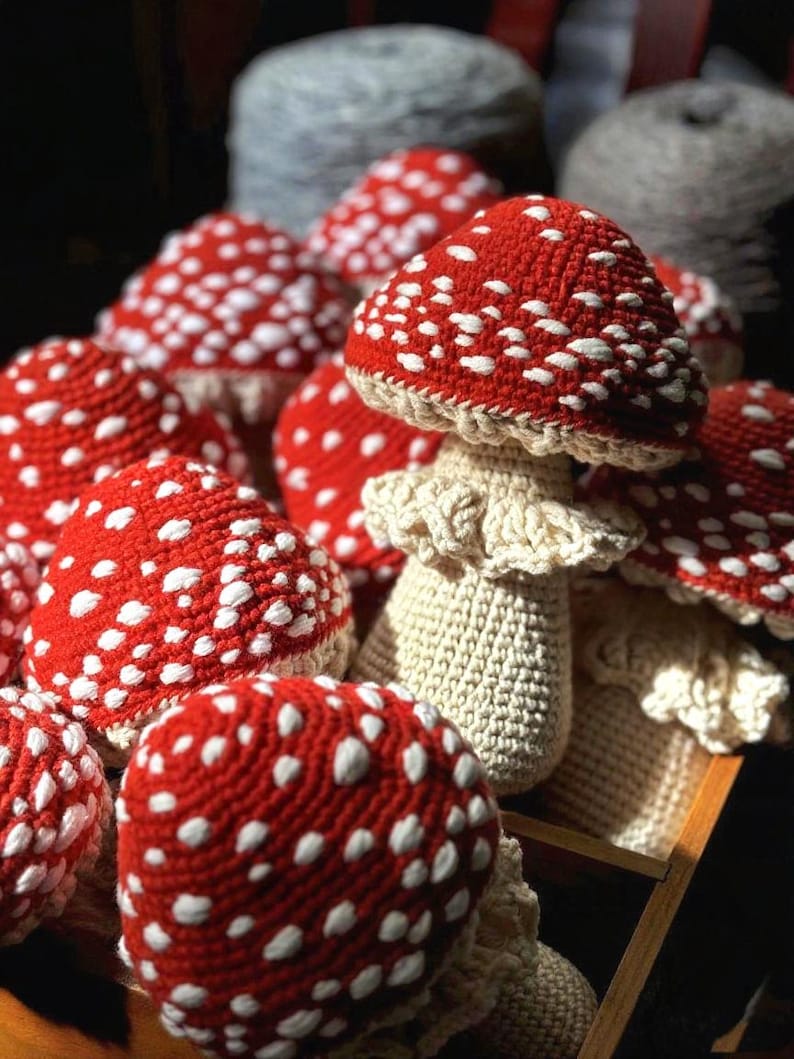 May include: A collection of handmade crochet mushrooms. The mushroom caps are red with white spots, and the stems are off-white. The mushrooms are arranged in a wooden tray, with some in the background. The yarn used appears to be a soft, textured material.