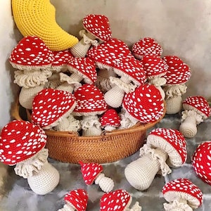 May include: A collection of handmade crochet mushrooms with red caps and white spots, displayed in a woven basket. A large yellow crescent moon is also present. The mushrooms vary in size and are arranged on a gray surface.