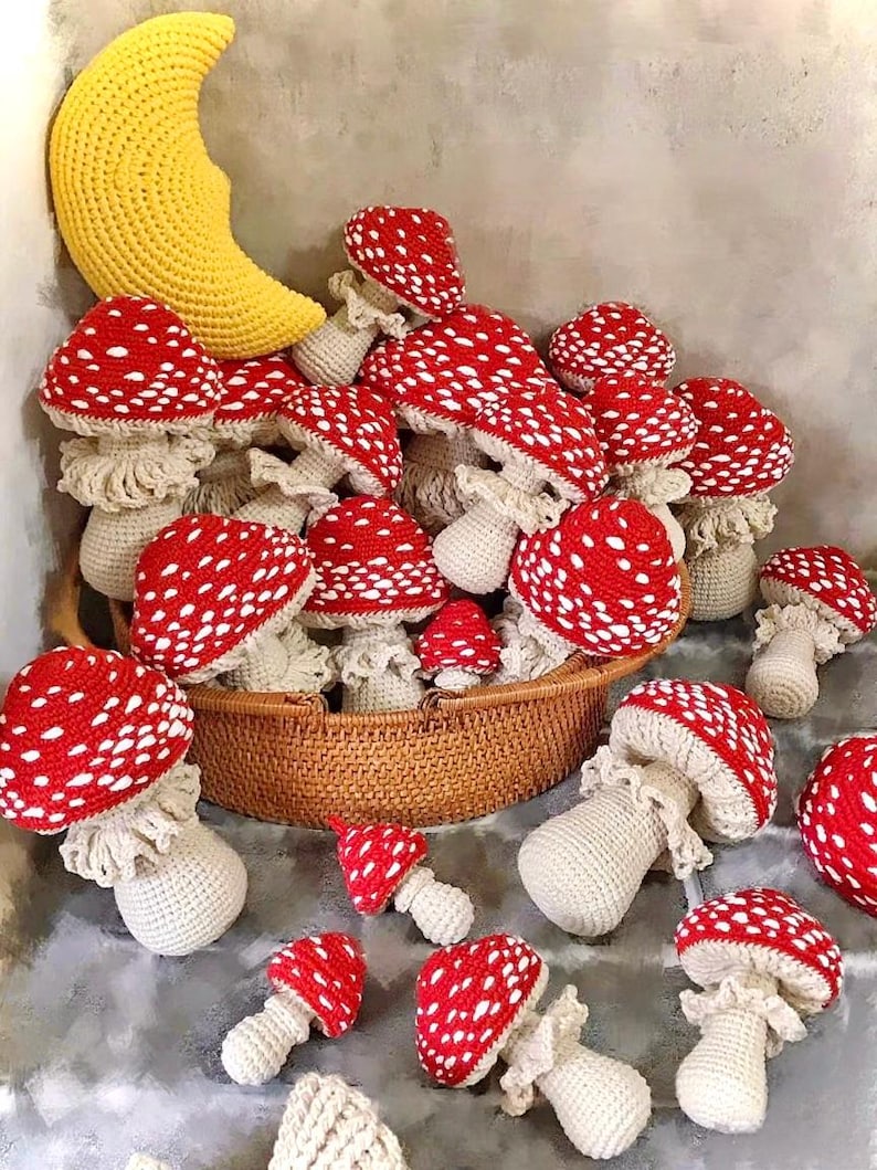 May include: A collection of handmade crochet mushrooms with red caps and white spots, displayed in a woven basket. A large yellow crescent moon is also present. The mushrooms vary in size and are arranged on a gray surface.