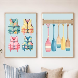 May include: Two framed wall art prints featuring nautical themes. The left print displays four colorful life vests in blue, yellow, pink, and floral patterns. The right print shows five wooden boat paddles in various shades hanging from a horizontal rack.