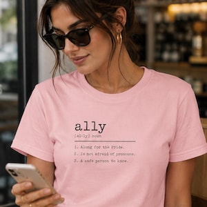 May include: A light pink short-sleeve t-shirt featuring black text that reads: ally [al-ly] noun 1. Along for the Pride. 2. Is not afraid of pronouns. 3. A safe person to know. The shirt has a crew neckline and a standard fit, worn with white pants.