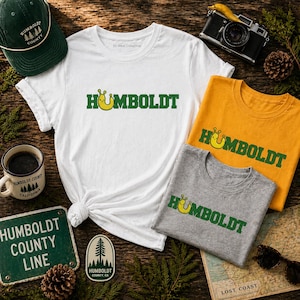 Puede incluir: Una colección de artículos con el tema del condado de Humboldt, que incluye camisetas blancas, amarillas y grises, una gorra verde, una taza de café y un letrero de metal. Las camisetas presentan la palabra "HUMBOLDT" en verde con un personaje de dibujos animados amarillo.