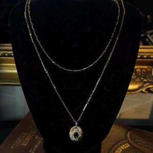 May include: A two-strand gold-toned necklace displayed on a black velvet bust. The top strand is a delicate chain, while the lower strand features a pendant with a dark stone center. The necklace is presented on a dark brown book.