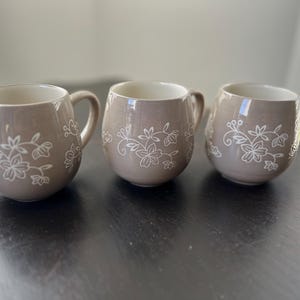 Könnte beinhalten: Drei taupefarbene Keramikbecher mit weißen Blumenmustern. Die Becher haben eine abgerundete Form mit Henkel und hellem Innenraum. Sie stehen auf einer dunklen Oberfläche.