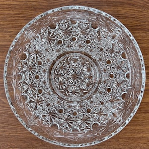 May include: Clear glass serving dish with a starburst pattern. The dish is divided into sections, with a central circular area and four surrounding compartments. The rim has a decorative edge. The dish is designed for serving snacks or appetizers.