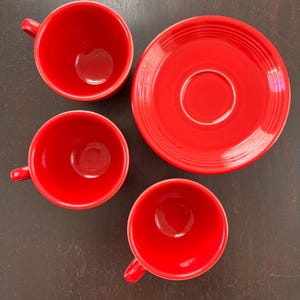 Puede incluir: Un juego de tres tazas de café rojas de cerámica con asas y un platillo a juego. Las tazas y el platillo son de un color rojo sólido y vibrante. Las tazas son redondas con un asa pequeña en el lateral. El platillo también es redondo con un centro rebajado.