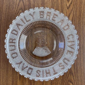 Op de afbeelding: Transparante glazen plaat met een geschulpte rand en de woorden "GIVE US THIS DAY OUR DAILY BREAD" in verhoogde letters. Het midden heeft een tarwe-schoof ontwerp. De plaat is rond en staat op een houten oppervlak.