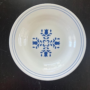 May include: A round white ceramic bowl featuring a blue floral pattern in the center and two thin blue circular lines running along the inner rim. The design consists of four stylized blue tulip-like flowers arranged in a cross shape.