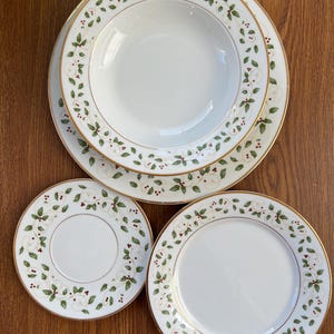 Op de afbeelding: Een set wit keramisch servies met een gouden rand en een hulstbessenontwerp. De set bevat een soepkom, twee dinerborden en een kleiner bord. Het hulstontwerp heeft groene bladeren en rode bessen.