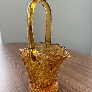 Könnte beinhalten: Bernsteinfarbener Glas-Korb mit Henkel. Der Korb hat ein Sternmuster und einen gewellten Rand. Der gewölbte Henkel ist an den Seiten befestigt. Das Glas hat einen warmen, goldenen Farbton. Dekorationsobjekt.