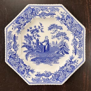 May include: An octagonal white plate with a blue floral border and a central blue pastoral scene. The scene depicts a person near a well, trees, and a village. The plate has a scalloped edge.