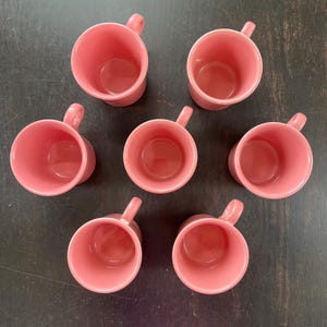 May include: Seven pink ceramic mugs with handles are arranged on a dark surface. The mugs are all the same size and shape, with a rounded body and a small handle. The interior of the mugs is also pink.
