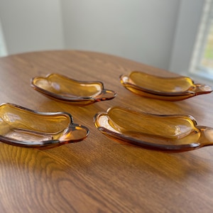 Pode incluir: Quatro travessas de vidro âmbar. Cada travessa tem um formato exclusivo, com bordas onduladas, base arredondada e borda alargada. As travessas estão dispostas sobre uma superfície de madeira, mostrando sua cor quente e translúcida e design.