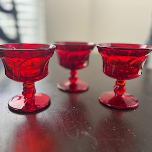 May include: Three matching ruby red glass dessert cups featuring a wide, flared rim and a short, decorative pedestal base. Each cup displays a translucent, deep red color with a smooth, polished finish and a sturdy, circular footed bottom.