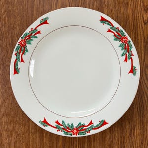 Puede incluir: Plato de cerámica blanca con borde dorado y decoraciones navideñas festivas. El plato presenta cintas rojas, agujas de pino verdes y flores de pascua rojas alrededor del borde, creando un tema navideño.