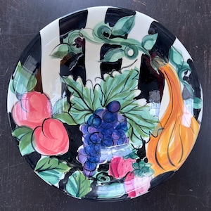 Puede incluir: Un plato decorativo de cerámica con un borde a rayas blancas y negras. El plato presenta un diseño colorido pintado a mano de frutas, incluyendo uvas moradas, melocotones y una calabaza naranja, con hojas y vides verdes.