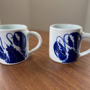 May include: Two white ceramic mugs with a blue lobster design. The mugs have a rounded shape with a handle and a light blue interior. The lobster design is painted in a bold, dark blue color.
