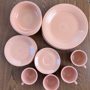 Puede incluir: Una colección de vajilla de cerámica rosa, que incluye platos, cuencos, platillos y tazas. Los platos y cuencos tienen detalles de anillos concéntricos. Las tazas tienen asas pequeñas. El juego está dispuesto sobre una superficie de madera.