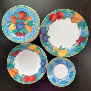 May include: A set of ceramic plates with a fruit design. The plates feature a white center surrounded by a blue and green border with illustrations of peaches, plums, lemons, and cherries. The set includes dinner plates, bowls, and saucers.