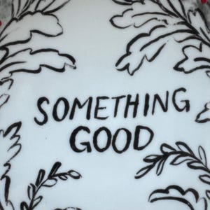 Puede incluir: Superficie blanca con las palabras "SOMETHING GOOD" en letras negras en negrita. El texto est&aacute; rodeado de dibujos lineales negros de hojas y ramas, creando un borde decorativo. El dise&ntilde;o es simple y elegante.