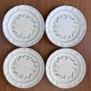 May include: Four white ceramic plates with a floral design. Each plate has a scalloped edge, a blue rim, and a wreath of colorful flowers in the center. The plates are arranged on a wooden surface.