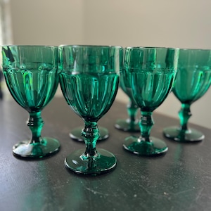 May include: A set of six emerald green wine glasses. Each glass features a ribbed bowl design and a detailed stem. The glasses are arranged on a dark surface, showcasing their elegant shape and color.