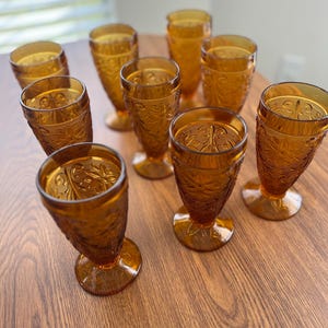 Könnte beinhalten: Eine Sammlung bernsteinfarbener Weingläser im Vintage-Stil. Jedes Glas weist ein geprägtes Blumendesign und einen abgerundeten Fuß auf. Die Gläser sind auf einer Holzoberfläche angeordnet, wodurch ihre Details und der warme Farbton hervorgehoben werden.