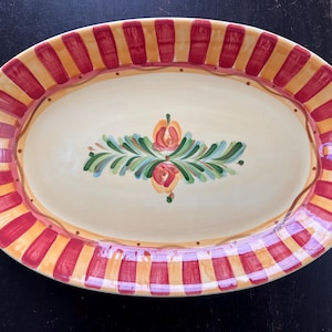 Op de afbeelding: Ovale serveerschaal met een crèmekleurig midden en een decoratieve rand. De rand heeft afwisselend rode en gele strepen. Het midden heeft een bloemenontwerp met groene bladeren en oranje bloemen. Een decoratief stuk voor het serveren van eten.