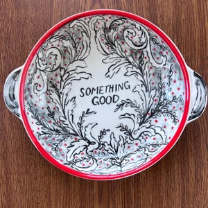 Puede incluir: Un cuenco de cer&aacute;mica blanca con borde y asas rojas. El cuenco presenta un dise&ntilde;o floral negro y gris con puntos rojos y las palabras "SOMETHING GOOD" en el centro. El cuenco est&aacute; sobre una superficie de madera.