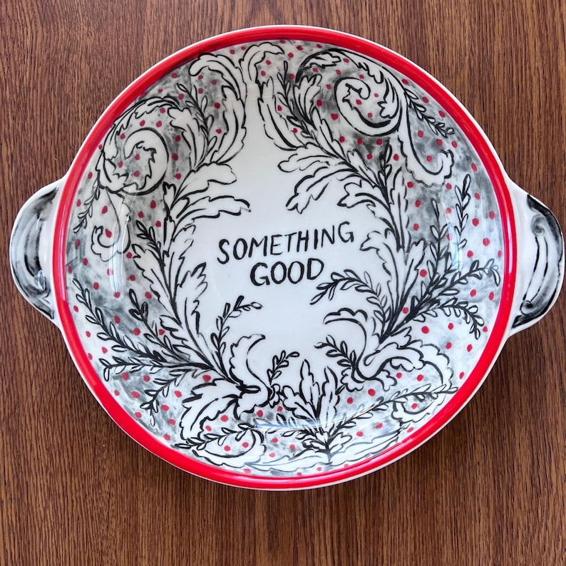 Puede incluir: Un cuenco de cer&aacute;mica blanca con borde y asas rojas. El cuenco presenta un dise&ntilde;o floral negro y gris con puntos rojos y las palabras "SOMETHING GOOD" en el centro. El cuenco est&aacute; sobre una superficie de madera.