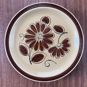 Puede incluir: Un plato de cerámica beige con un diseño floral marrón. El plato presenta dos flores marrones con contornos negros y hojas marrones. El borde tiene una doble raya marrón. El plato está sobre una superficie de madera.