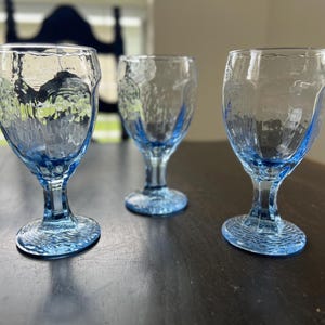 Pode incluir: Três taças de vidro azul-claro com uma superfície texturizada e ondulada. Cada taça apresenta uma haste curta e robusta e uma base circular com um padrão em relevo. O vidro tem uma qualidade translúcida com um tom azul distinto.