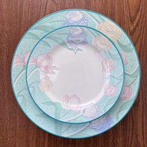 Puede incluir: Dos platos redondos de cerámica con un diseño floral. Los platos tienen un fondo verde azulado claro con un borde turquesa. El diseño floral incluye tulipanes rosas, amarillos y azules con hojas verdes.