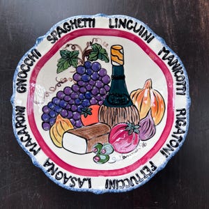 Puede incluir: Un plato decorativo de cerámica con borde festoneado, que presenta una ilustración central de uvas, una botella de vino y varias verduras. El plato está rodeado por los nombres de platos de pasta en letras negras, con un borde rojo y blanco.