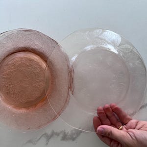 May include: Two pink glass plates with a floral pattern. One plate is a darker pink, and the other is a lighter, almost clear pink. The plates are round with a slightly raised edge. The plates are likely for serving or display.