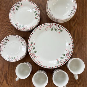 Könnte beinhalten: Ein weißes Weihnachtsgeschirr-Set mit roten und grünen Stechpalmen- und Schleifenakzenten. Das Set enthält Teller, Schalen, Untertassen und Tassen. Die Ränder der Teller und Schalen haben einen rot-grünen gepunkteten Rand.