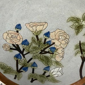 Puede incluir: Un plato de cerámica con fondo azul claro y borde marrón. El plato está decorado con un diseño floral con rosas color crema, capullos azules y hojas verdes. El plato redondo parece hecho a mano.