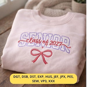 May include: A light pink sweatshirt with the embroidered text "SENIOR class of 2027" in purple and red. A red bow is embroidered below the text. The sweatshirt is folded on a wooden surface.