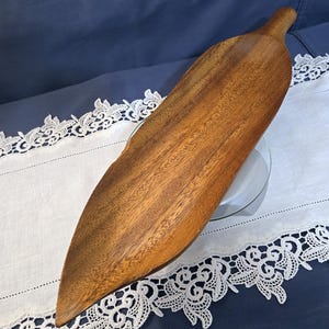 Puede incluir: Una bandeja de madera con forma de hoja, con un acabado de veta de madera natural. La bandeja tiene un asa y está colocada sobre un paño blanco con ribete de encaje sobre un fondo azul. Esta bandeja decorativa es adecuada para servir alimentos.