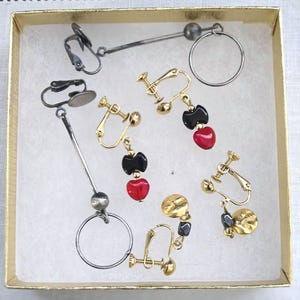 May include: A collection of clip-on earrings in a gold box. The earrings feature various designs, including gold-toned metal, black and red heart-shaped beads, and circular elements. Some earrings have a long, linear design.
