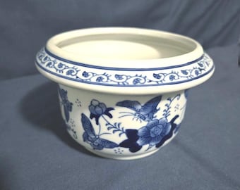 Puede incluir: Una maceta de cerámica blanca y azul con un borde ancho. La maceta presenta un diseño floral y de mariposas azul. El borde tiene un patrón decorativo azul. La maceta es redonda y tiene una amplia abertura.