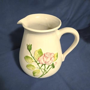 May include: White ceramic pitcher with a curved handle and spout. The pitcher is decorated with a pink rose and green leaves. The pitcher is likely used for serving beverages.