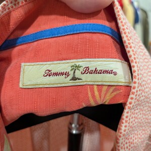 May include: Close-up of a coral-colored button-down shirt with a blue collar lining. The shirt features a label with the text "Tommy Bahama" and a palm tree logo. The fabric has a subtle textured pattern and a white dotted design.