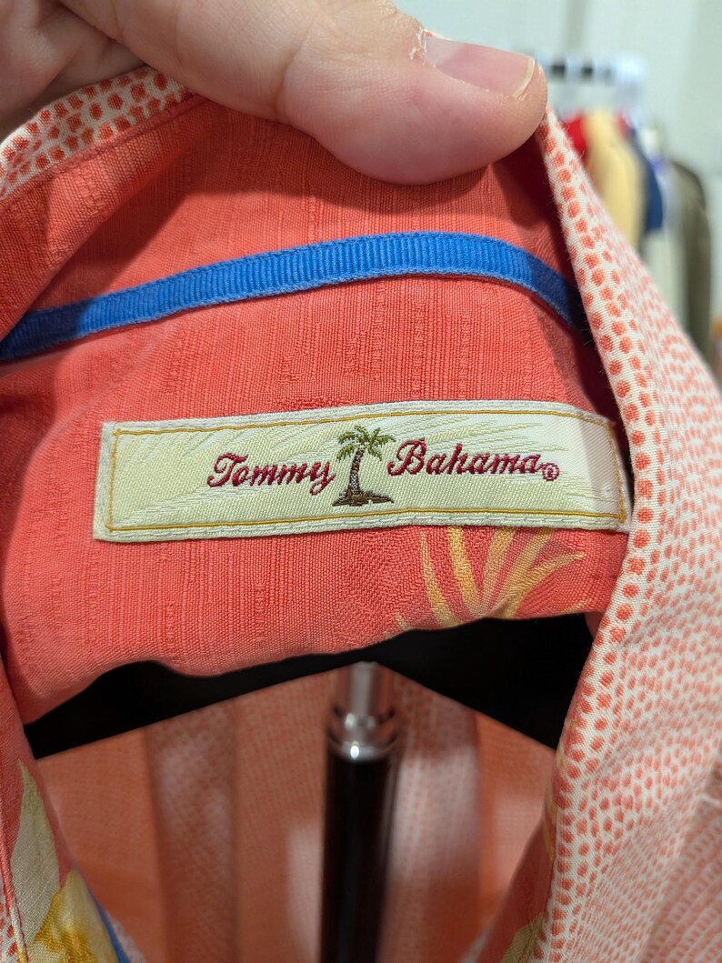 May include: Close-up of a coral-colored button-down shirt with a blue collar lining. The shirt features a label with the text "Tommy Bahama" and a palm tree logo. The fabric has a subtle textured pattern and a white dotted design.